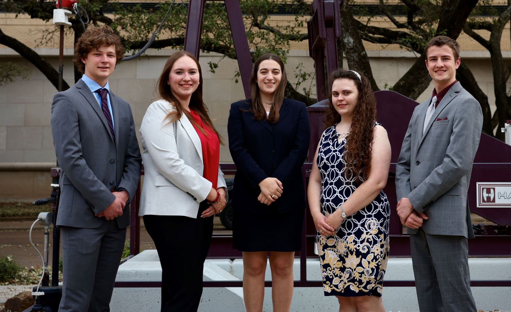 Five students dressed in business attire standing in front of a large maroon structure, smiling at the camera.