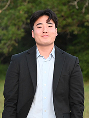 Kim wears a black suit jacket and blue button-down shirt, in front of a grove of trees.