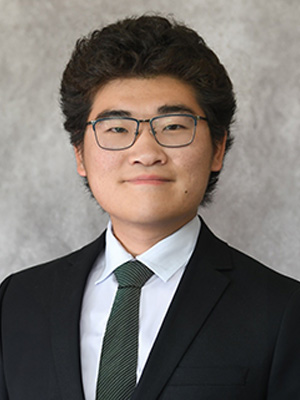 Kwon wears a black suit, green tie and glasses, smiling, standing in front of a grey backdrop.