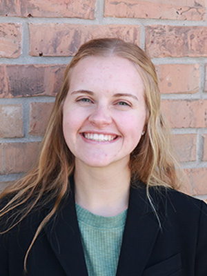 Headshot of Megan in front of a brick wall, wearing a dark suit jacket and blue shirt.