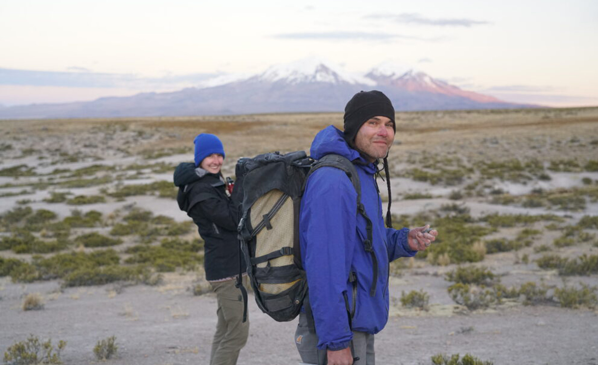 Two hikers with backpacks smiling at the camera in a vast landscape, with a mountain in the background during dusk.