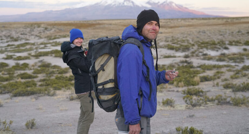 Two hikers with backpacks smiling at the camera in a vast landscape, with a mountain in the background during dusk.