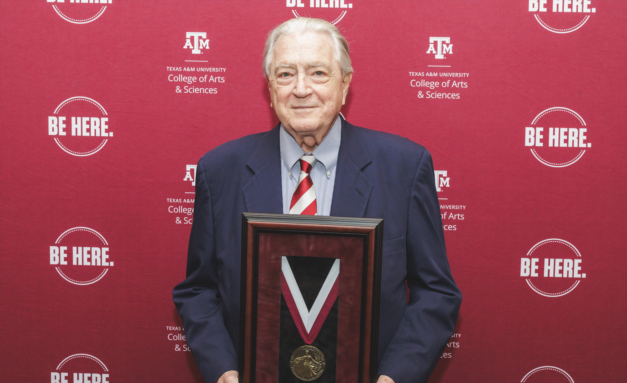 Luke Soules stands in front of a maroon back drop with the logo that says "Be Here," holding his Medal for Social Sciences at the 2025 Spring Banquet.