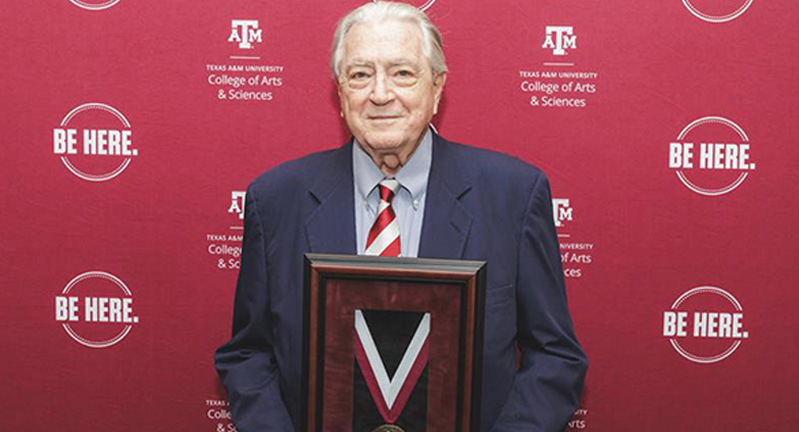 Luke Soules stands in front of a maroon back drop with the logo that says "Be Here," holding his Medal for Social Sciences at the 2025 Spring Banquet.