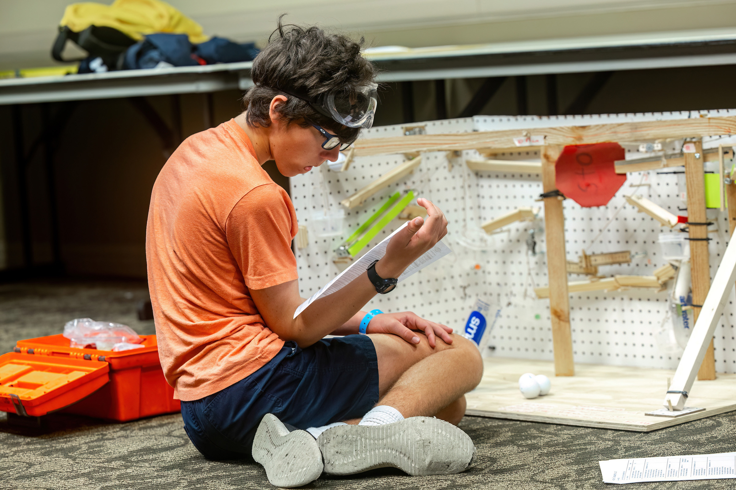 Participant at the Science Olympiad sits on the floor, working on a robotic component with tools and materials around them.