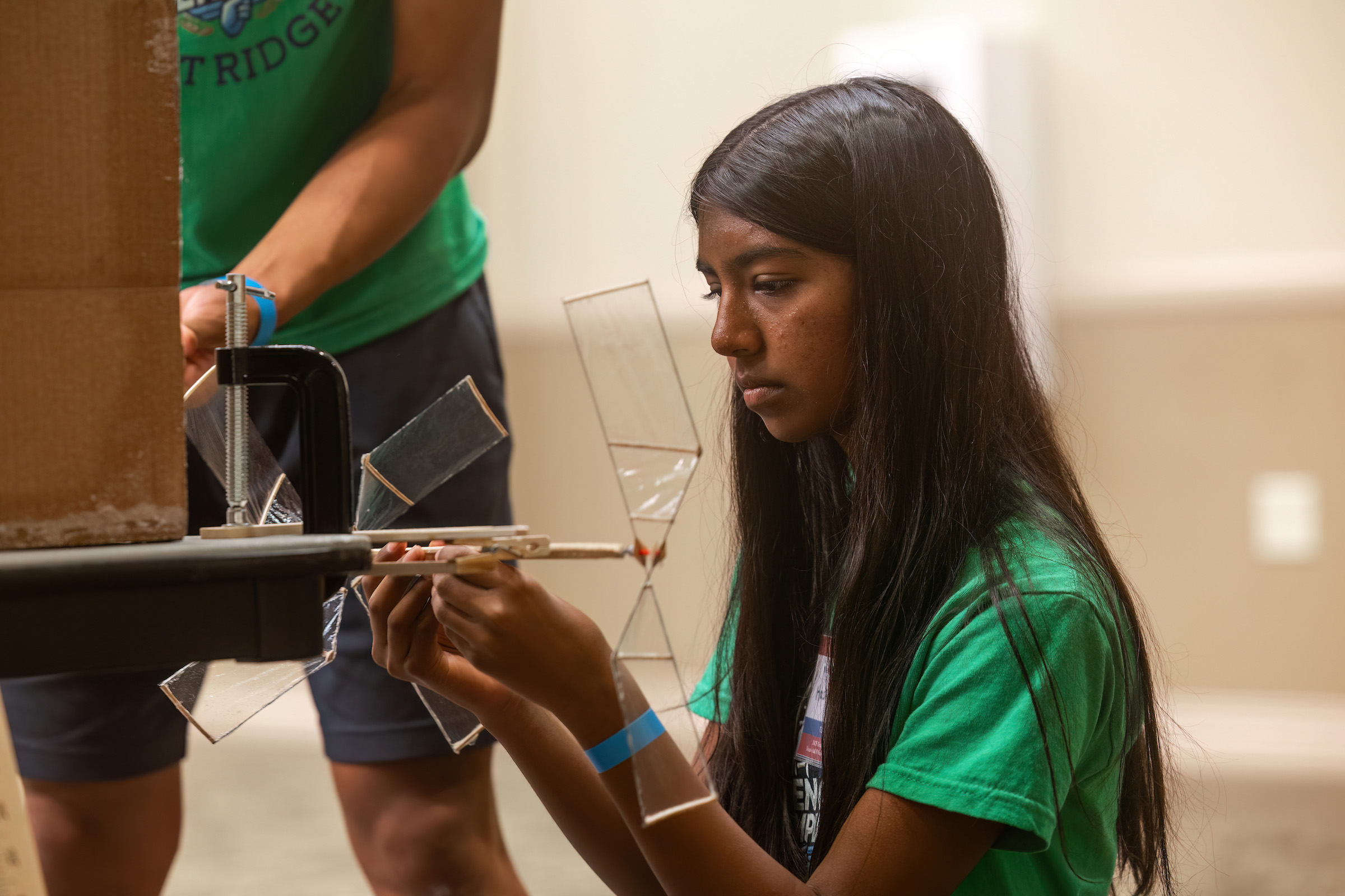 Person in a green T-shirt focusing intently on assembling a small, intricate model at a workshop. Another participant is partially visible in the background.