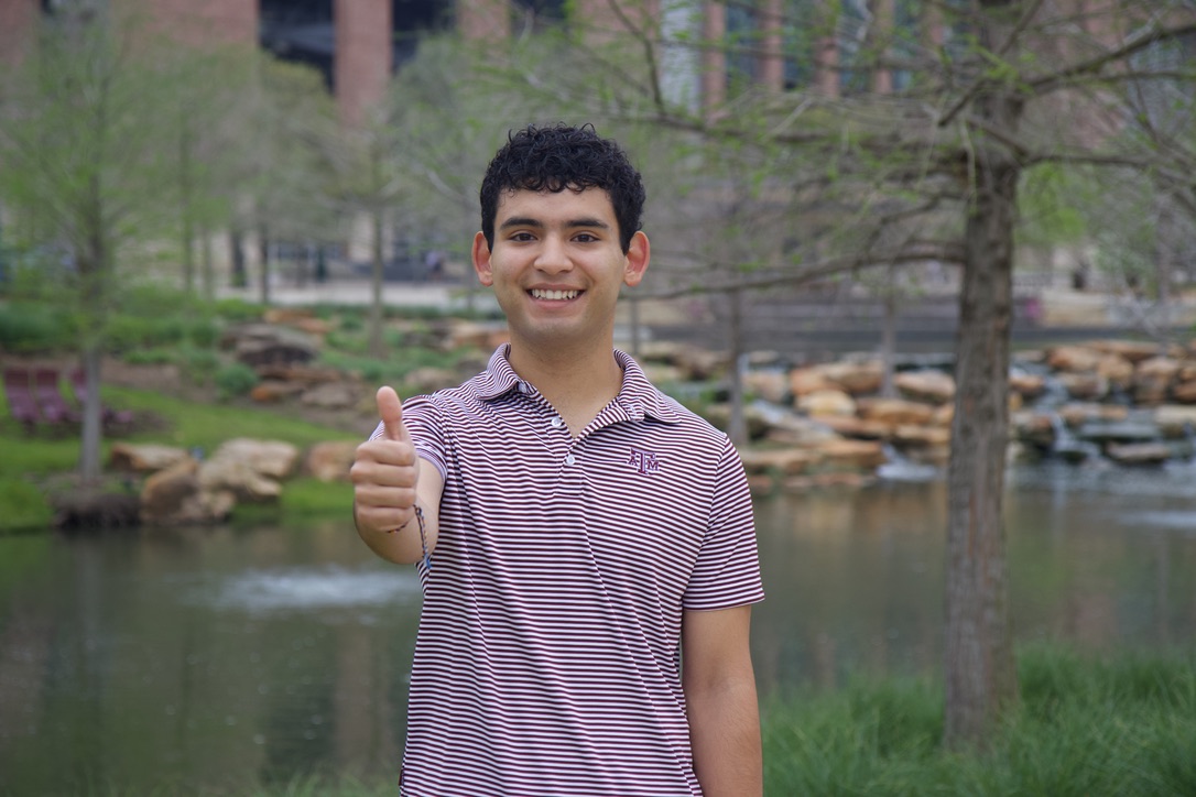 Person giving a thumbs up in a park with a pond and trees in the background.