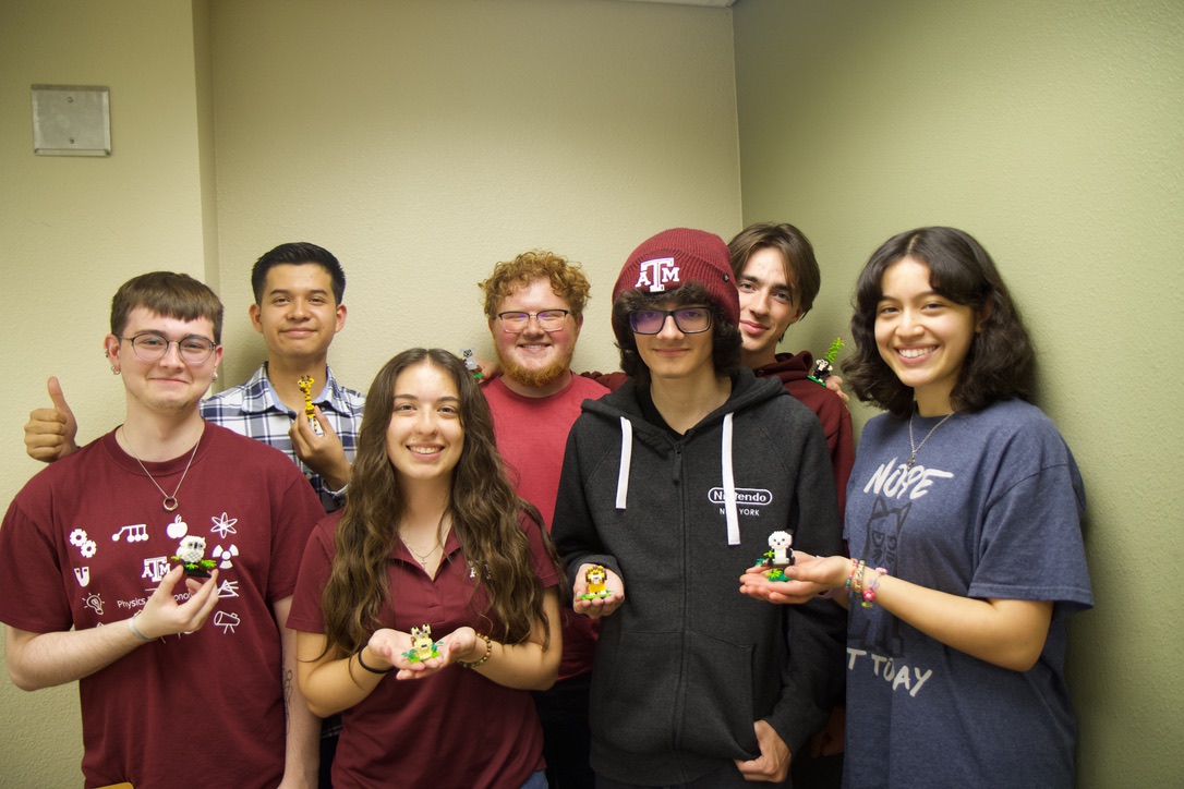 Group of seven individuals in a room, smiling and holding colorful puzzle cubes. Some are wearing shirts with logos including Texas A&M and Google.