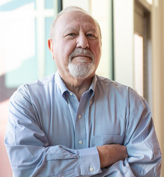 Ersen Arseven poses in front of an open window in a blue shirt with arms crossed and a small smile.