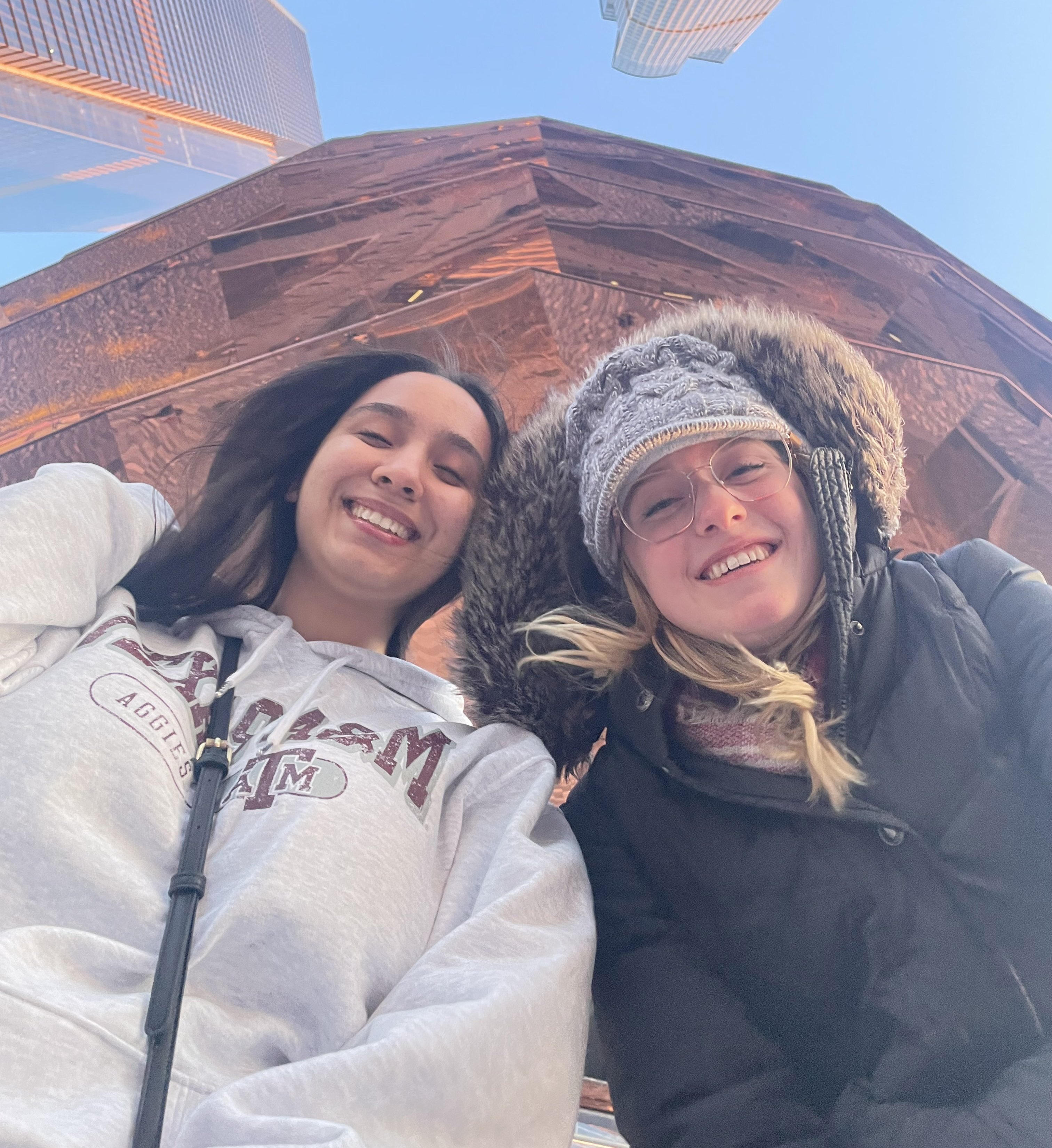 Two people smiling in front of the Vessel on a sunny day, one wearing a knit hat and the other in a hoodie.