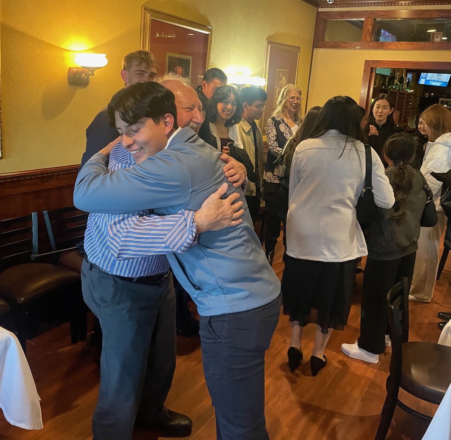 Two people embracing in a warm hug at a social gathering in a restaurant setting, with other guests chatting in the background.