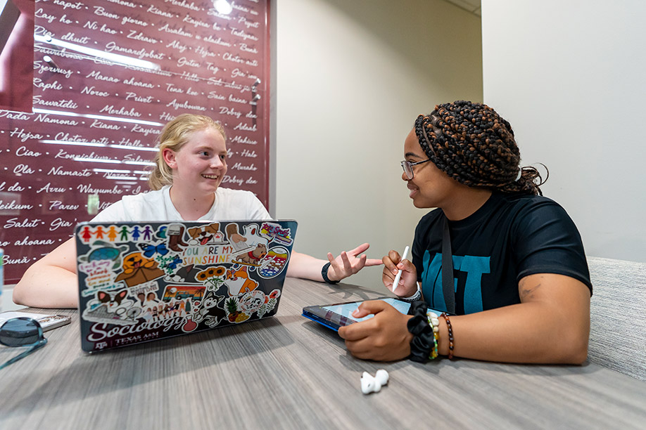 two college girls sitting at desk with laptops and tablets discussing an assigment